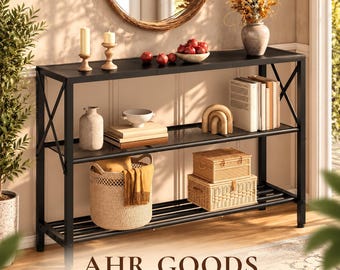 May include: A black metal console table with three tiers, featuring X-shaped side supports. The shelves hold a woven basket, two lidded wicker boxes, books, a ceramic vase, candles, a bowl of red fruit, and a decorative wooden arch.