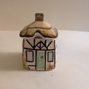 May include: A ceramic cottage-shaped jar with a removable thatched roof lid. The structure features white walls with black timber framing, a central light green door, two small windows, and painted floral details on the sides.