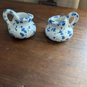 May include: Two small ceramic creamers featuring a white base with a blue speckled pattern. Each piece has a rounded body, a short spout, and a single curved handle attached to the side. The glaze is smooth and glossy, showing irregular blue spots.