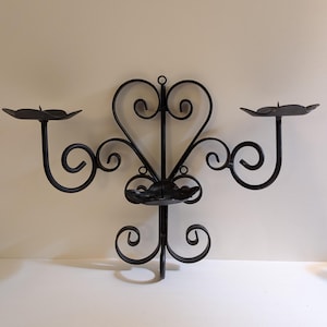 May include: A black metal wall-mounted candle holder featuring three circular drip plates with central spikes. The frame is constructed from curved iron rods arranged in symmetrical scrollwork patterns, including a heart-shaped center element.
