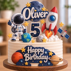 May include: A round cake decorated with space-themed toppers including an astronaut, a rocket ship, a satellite, planets, and stars. The cake features the name Oliver, the number 5, and the text Happy Birthday in blue and white lettering.