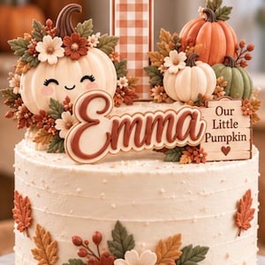 May include: A round white cake decorated with autumn-themed fondant elements including a large number one with a brown gingham pattern, pumpkins, fall leaves, and flowers. The cake features the name Emma and a sign reading Our Little Pumpkin.
