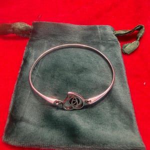 Könnte beinhalten: Ein silberfarbener Metallarmreif mit einem zentralen herzförmigen Anhänger, der ein filigranes, dunkel ausgefülltes Ausschnittmuster aufweist. Das Armband hat eine glatte, polierte Oberfläche und liegt auf einem dunkelgrünen Samtbeutel.