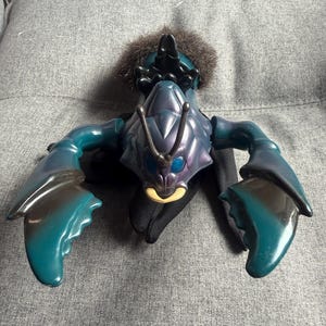 May include: A plastic action figure resembling a crustacean with a dark teal and purple shell, large articulated claws, blue eyes, and a tan mouth area. The figure features a tuft of brown faux fur on its back and black fabric cape-like wings.