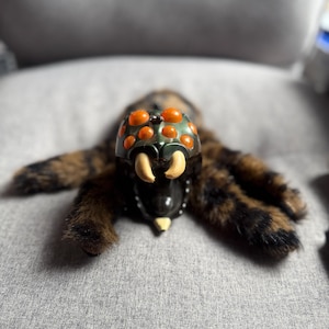May include: A plush spider toy featuring a dark green plastic head with eight bright orange circular eyes and two curved cream-colored fangs. The body is covered in long, soft brown and black faux fur, extending into multiple articulated legs.