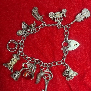 May include: A silver charm bracelet featuring various dangling charms including a violin, a heart-shaped padlock, a key, a clock, a bell, a shoe, a carriage, and a small sphere with a red stone. The bracelet consists of a linked silver chain.