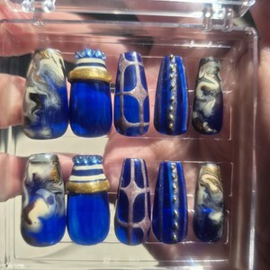 Puede incluir: Un juego de diez uñas postizas en un estuche de plástico transparente, con una paleta de colores azul intenso. Los diseños incluyen remolinos de mármol azul y blanco con detalles dorados, uñas azul sólido con bandas doradas, patrones de cuadrícula y detalles de tachuelas doradas verticales.