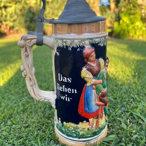May include: A ceramic beer stein with a dark blue body, a metal lid, and a decorative handle. The stein features a relief illustration of two figures in traditional clothing and the German text "Das lieben wir" printed in white on the side.