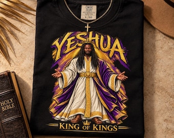 May include: A black t-shirt featuring a graphic of a figure with long dark hair wearing a crown, a white robe with gold trim, and a purple cloak. The text YESHUA appears above the figure, and KING OF KINGS is printed below in gold lettering.