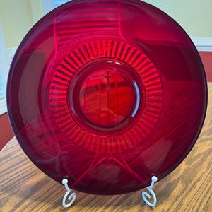May include: A circular, translucent ruby red glass plate featuring a central medallion design surrounded by a radiating sunburst pattern of vertical ridges. The plate is displayed upright on a white wire stand.