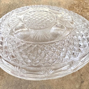 May include: A clear glass bowl with a lid featuring a textured diamond pattern across the surface. The lid has a central circular medallion with a cross-hatched design, while the base and lid edges are decorated with a radiating fan pattern.