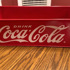 May include: A rectangular red metal storage bin featuring the white text Drink Coca-Cola on the front side. The container has an open top and a smooth finish, displaying the iconic brand logo prominently across the center of the exterior.