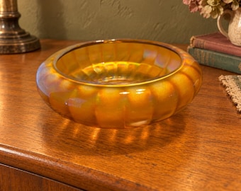 May include: A round, amber-colored glass bowl featuring a ribbed, scalloped exterior design with an iridescent finish that reflects light, creating a shimmering effect across the surface of the translucent, circular vessel.