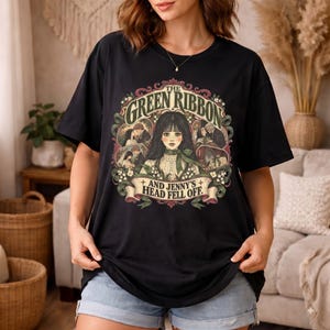 以下が含まれることがあります： THE GREEN RIBBON AND JENNY'S HEAD FELL OFFというテキストがプリントされた黒の半袖Tシャツ。中央の人物のイラストの周りに、小さなシーン、花のアクセント、装飾的なリボンが描かれたグラフィックデザインが特徴です。