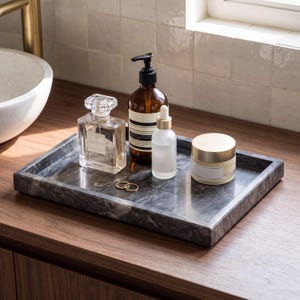 May include: A rectangular dark gray marble tray with white veining holding a clear glass perfume bottle, a brown amber pump bottle, a small frosted glass dropper bottle, a round cream jar with a gold lid, and two thin gold metal rings.
