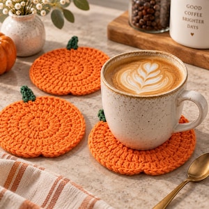 Puede incluir: Tres posavasos redondos de ganchillo de color naranja con forma de calabaza y pequeños tallos verdes. Un posavasos sostiene una taza de cerámica moteada llena de café con arte latte sobre una superficie plana.