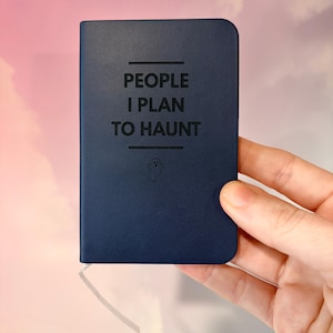 Könnte beinhalten: Ein dunkelblaues Taschennotizbuch mit abgerundeten Ecken. Der Einband zeigt den schwarzen Text "PEOPLE I PLAN TO HAUNT", der zwischen zwei horizontalen Linien zentriert ist, mit einem kleinen, einfachen Geistersymbol unter dem Text.
