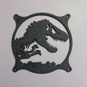 May include: A black, circular fan grill featuring a cutout silhouette of a Tyrannosaurus Rex dinosaur. The frame has four mounting holes at the corners and a textured finish, showcasing the dinosaur's head, teeth, and small arms in profile.