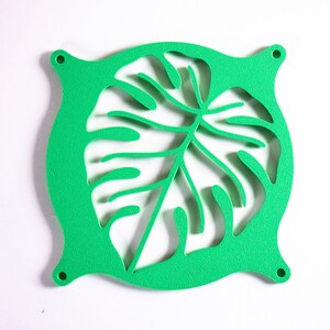 May include: A bright green square fan grill featuring a laser-cut monstera leaf design in the center. The frame has rounded edges with a small circular mounting hole at each of the four corners, providing a decorative and functional cover.