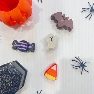 May include: A collection of Halloween-themed items including an orange pumpkin container, a brown bat, a white ghost, a black and purple candy, a candy corn piece, a black hexagonal glitter container, and several black plastic spider decorations.