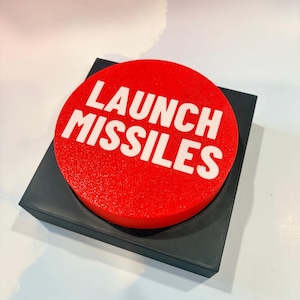 May include: A circular red button with the white text LAUNCH MISSILES printed on the top surface. The button is mounted on a square black base with a textured finish. The design features a bold, high-contrast aesthetic with clear lettering.