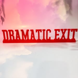 May include: A bright red, free-standing sign featuring the words DRAMATIC EXIT in a bold, block-style font. The letters are connected at the base, creating a single, solid, horizontal piece of decor that stands upright on a flat surface.