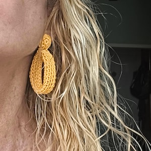Op de afbeelding: Een enkele gele gehaakte oorbel in een druppelvorm. De oorbel is gemaakt van een geweven, getextureerd materiaal. De oorbel wordt gedragen aan het oor van een persoon met lang, golvend blond haar.