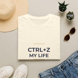 Puede incluir: Una camiseta de color crema con el texto azul oscuro CTRL+Z MY LIFE impreso en la parte delantera. La prenda tiene un cuello redondo y una textura de tela moteada, presentada en plano con un diseño sencillo y limpio.