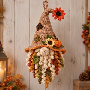 Puede incluir: Un adorno de pared de ganchillo con forma de gnomo que presenta un sombrero puntiagudo de color tostado decorado con un girasol naranja brillante, una pequeña calabaza naranja y hojas verdes. El gnomo tiene una nariz redonda beige y una barba larga y texturizada hecha de hilo color crema, naranja y verde.