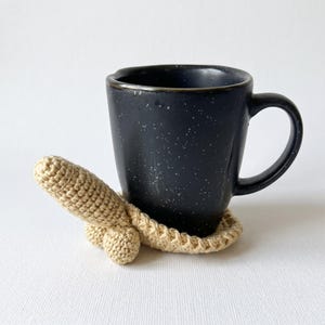 Può includere: Una tazza in ceramica nera con macchie bianche poggia su un sottobicchiere all'uncinetto color beige. Il sottobicchiere presenta una base circolare con un elemento tridimensionale a forma fallica che sporge dal bordo anteriore.
