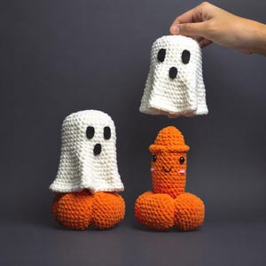 Op de afbeelding: Twee witte gehaakte spookfiguren met zwarte vilten ogen en monden, de ene geplaatst op een oranje gehaakte pompoenbasis en de andere omhoog gehouden, naast een oranje gehaakte fallische figuur met een glimlachend gezicht en roze wangen.