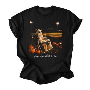 May include: A black t-shirt featuring a graphic of a ghost sitting in a folding chair on a baseball field at night. The ghost holds a steaming cup, surrounded by orange pumpkins and string lights. The text "yup, i'm still here" is printed below.