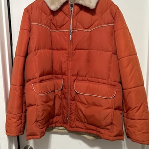 May include: A quilted orange jacket featuring a cream-colored faux fur collar, a full-length silver-tone front zipper, and two front patch pockets with cream piping. The jacket has a boxy silhouette with horizontal and vertical stitching patterns.