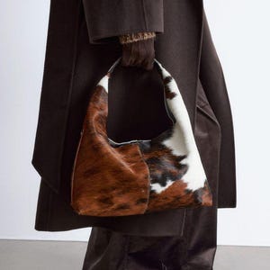 May include: A hobo-style handbag crafted from cowhide featuring a distinct tri-color pattern of brown, black, and white patches. The bag has a single integrated handle and a curved silhouette, showcasing the natural texture of the animal hide.