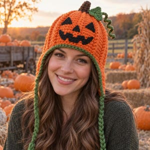 May include: A bright orange crocheted beanie designed to look like a jack-o'-lantern, featuring a black stitched face, a brown stem, a green leaf, and a curly green vine. The hat includes long, braided green earflaps hanging down on both sides.