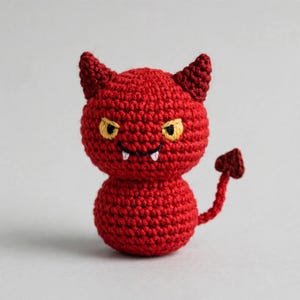Puede incluir: Una pequeña criatura roja tejida a ganchillo con dos cuernos puntiagudos, ojos amarillos y dos colmillos blancos. La figura tiene cabeza y cuerpo redondos, y una cola delgada que termina en una punta con forma de corazón. Textura de hilo.