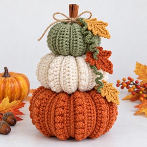 Peut inclure: Une décoration de citrouilles empilées au crochet comprenant trois citrouilles vert sauge, crème et orange brûlé. La citrouille supérieure est surmontée d'un bâton de cannelle et d'un nœud en ficelle, avec des feuilles crochetées orange et jaune.
