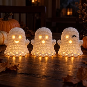 Puede incluir: Tres figuras de fantasmas de ganchillo blanco con bordes inferiores festoneados. Cada fantasma presenta un interior hueco iluminado por una luz amarilla cálida, con expresiones faciales recortadas que incluyen ojos y bocas cuadradas.