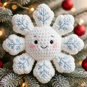 May include: A white crocheted snowflake ornament with a plush, rounded center featuring black embroidered eyes, a small smile, and pink blush cheeks. Each of the six points is decorated with light blue stitched snowflake patterns.