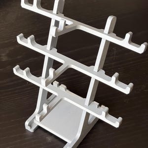 Può includere: Un espositore a gradini di colore grigio chiaro dotato di tre barre orizzontali con molteplici ganci a forma di U per organizzare oggetti. La struttura è sostenuta da due pannelli laterali verticali collegati a una base rettangolare piatta.