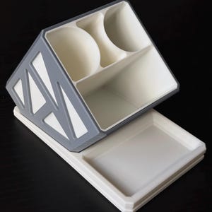 May include: A desk organizer featuring a white base with a rectangular tray and an angled upper section. The upper part has three compartments and is framed in gray with a geometric cutout pattern on the side, providing storage for stationery items.
