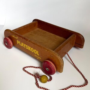 May include: A vintage wooden toy wagon with a brown finish, featuring red wheels and yellow text that reads "PLAYSKOOL wagon" and "colorol" on the side. A red and white braided pull cord with a small yellow wooden bead is attached to the front.
