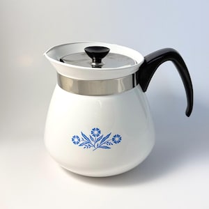 May include: A white teapot with a rounded body featuring a blue floral pattern on the front. It has a black handle, a stainless steel band around the upper section, and a matching lid with a small black knob handle on top for easy lifting.