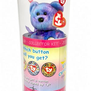 May include: A Ty Beanie Baby bear with purple and blue tie-dye fur inside a clear plastic collector kit container. The packaging features the text "Which button will you get?" and "Join the club!" with images of circular buttons and a red Ty heart tag.