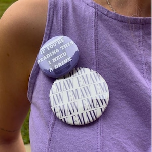 May include: Two circular pin-back buttons attached to a lavender sleeveless top. The smaller button is light purple with the white text IF YOU'RE READING THIS I NEED A DRINK. The larger button is white with the repeating dark text EMAW.