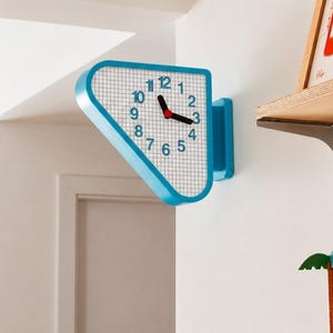 Pode incluir: Um relógio de parede com uma moldura triangular azul brilhante e um mostrador branco com um padrão de grade preta. O relógio exibe algarismos arábicos azuis de 1 a 12, ponteiros pretos de horas e minutos e um pequeno centro vermelho.