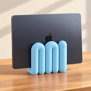 Puede incluir: Un portátil gris oscuro con un logotipo de Apple negro se mantiene en posición vertical mediante un soporte tubular en forma de U de color azul claro. El soporte presenta un diseño serpenteante, continuo y redondeado que sostiene el dispositivo.