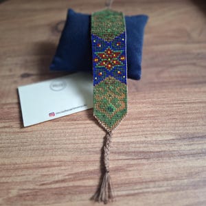 May include: A woven beaded bracelet featuring a geometric pattern in shades of green, blue, red, and yellow. The bracelet has a pointed end and a braided brown cord tie with a tassel. The design includes a central star motif and intricate beadwork.