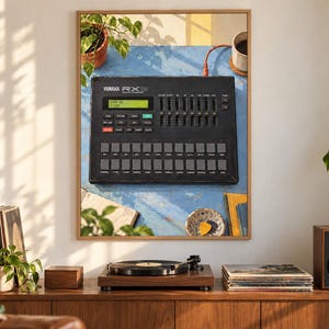 Pode incluir: Uma impress&atilde;o de arte de parede emoldurada apresentando uma bateria eletr&ocirc;nica Yamaha RX preta com display digital, v&aacute;rios controles deslizantes e uma grade de bot&otilde;es quadrados. O dispositivo est&aacute; sobre uma superf&iacute;cie azul com planta e caneca.