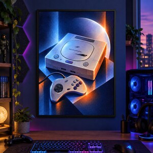 May include: A framed wall art print featuring a Sega Saturn game console and its matching controller. The console is light grey with the Sega Saturn logo on the disc lid. The artwork uses a high-contrast lighting style with blue and orange tones.